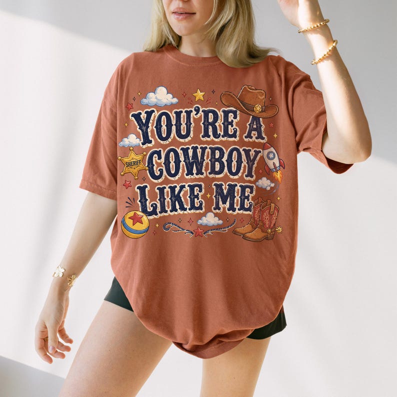 May include: A rust-colored t-shirt featuring the text "YOU'RE A COWBOY LIKE ME" in dark blue lettering. The graphic includes a brown cowboy hat, a sheriff star badge, a rocket ship, a colorful ball, and cowboy boots, surrounded by small clouds and stars.