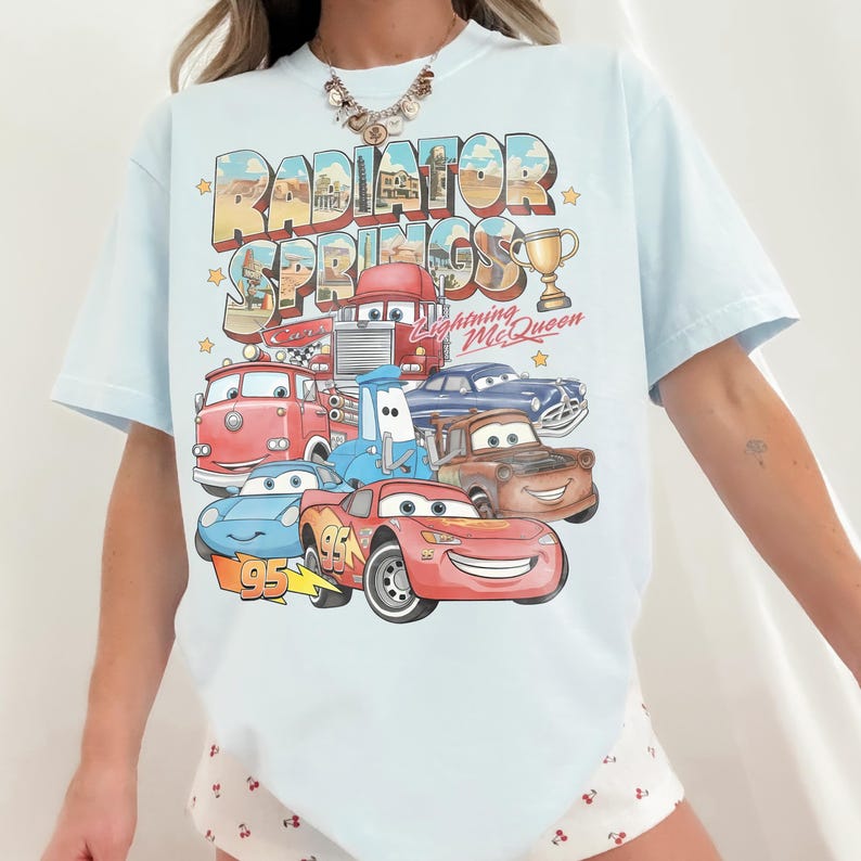Op de afbeelding: Een lichtblauw t-shirt met een grafische print van personages uit de film Cars, waaronder Lightning McQueen, Takel en anderen. De tekst Radiator Springs, Cars en Lightning McQueen staat gedrukt boven de kleurrijke illustraties van voertuigen.