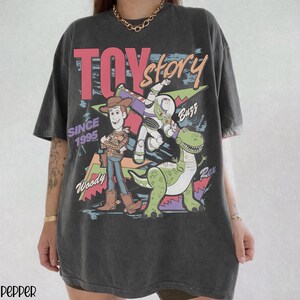 Pode incluir: T-shirt cinzento escuro com um gr&aacute;fico colorido de Toy Story com Woody, Buzz Lightyear e Rex. O design inclui o texto "TOY STORY" e "SINCE 1995". A t-shirt tem um aspeto vintage e desgastado.