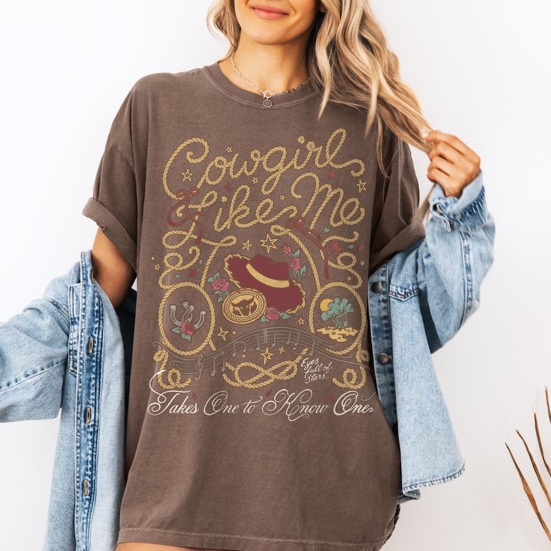 May include: A dark brown short-sleeve t-shirt featuring a graphic print with the text Cowgirl like Me and Takes One to Know One. The design includes a cowboy hat, a horseshoe, a rope border, musical notes, and small floral accents.
