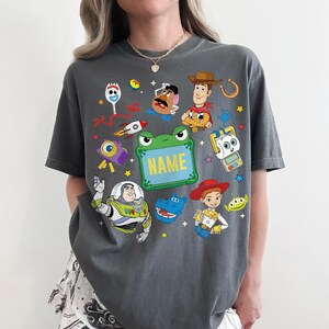 May include: A charcoal gray t-shirt featuring a colorful graphic print of various animated characters including a cowboy, a space ranger, a cowgirl, a potato-shaped figure, a fork-like character, and a green frog-shaped frame with the text NAME.