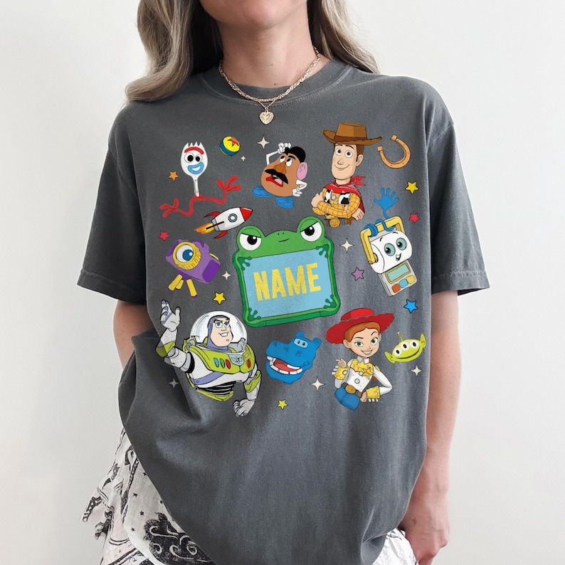 May include: A charcoal gray t-shirt featuring a colorful graphic print of various animated characters including a cowboy, a space ranger, a cowgirl, a potato-shaped figure, a fork-like character, and a green frog-shaped frame with the text NAME.