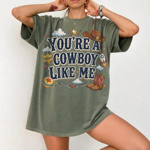 May include: An olive green short-sleeve t-shirt featuring the text "YOU'RE A COWBOY LIKE ME" in dark blue lettering. The graphic includes a brown cowboy hat, a yellow sheriff star badge, a rocket ship, a red and yellow ball, and brown cowboy boots.