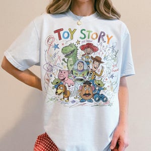 May include: Light blue t-shirt featuring a colorful, hand-drawn style graphic of Toy Story characters. The design includes the words "TOY STORY" in a playful font, along with images of Woody, Buzz Lightyear, and other characters from the movie.