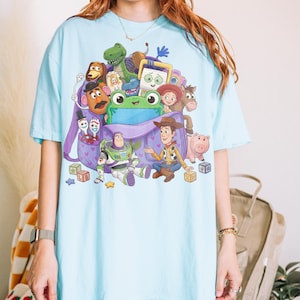 May include: Light blue t-shirt featuring a colorful graphic of characters from the animated film "Toy Story." The design includes Woody, Buzz Lightyear, and other characters, along with a purple backpack. The shirt is a casual, short-sleeved style.