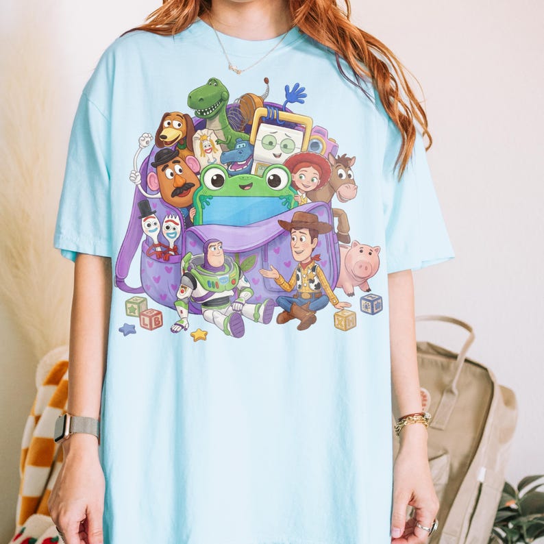 May include: Light blue t-shirt featuring a colorful graphic of characters from the animated film "Toy Story." The design includes Woody, Buzz Lightyear, and other characters, along with a purple backpack. The shirt is a casual, short-sleeved style.