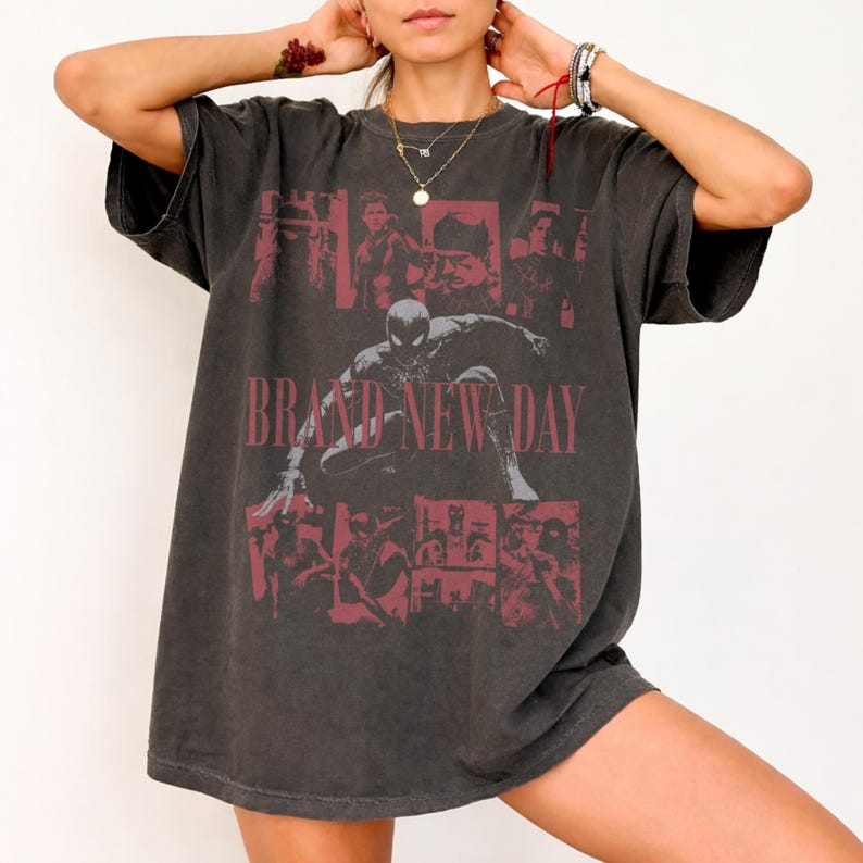 May include: A charcoal grey short-sleeve t-shirt featuring a faded red graphic print of a superhero character in a crouching pose. The design includes multiple smaller character panels and the text BRAND NEW DAY in a bold, distressed red font.