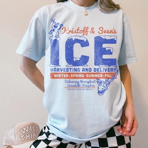 May include: Light blue t-shirt featuring the text "Kristoff & Sven's ICE" in blue and red, with a distressed vintage design. The shirt also includes illustrations of Olaf and Sven, and the text "Harvesting and Delivery".