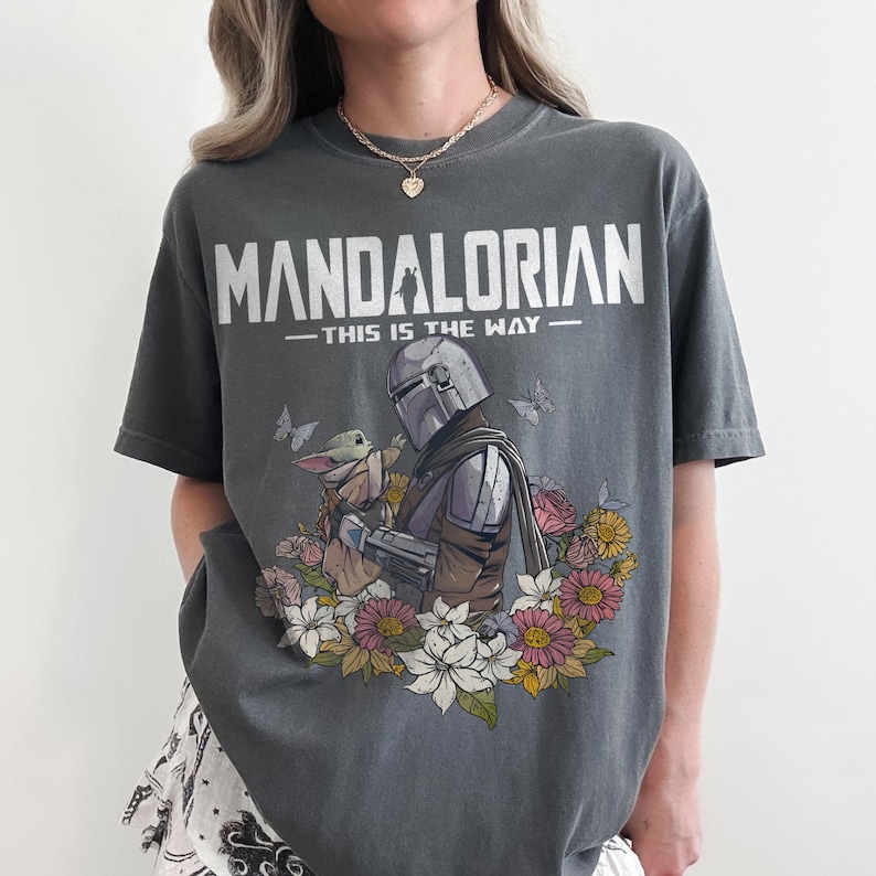 May include: Dark gray t-shirt featuring the text "MANDALORIAN" above "THIS IS THE WAY." The shirt displays an illustration of the Mandalorian and Grogu surrounded by flowers and butterflies. A casual, unisex tee.