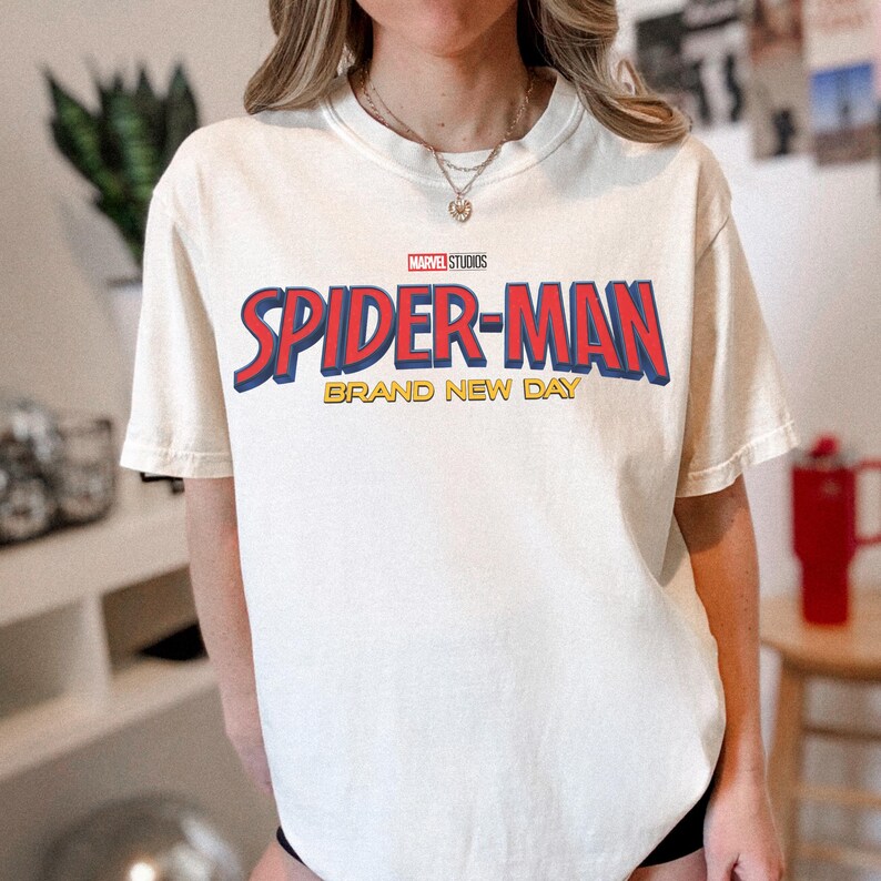 May include: A cream-colored short-sleeve t-shirt featuring the red and blue Marvel Studios Spider-Man logo with the text Brand New Day in yellow underneath. The shirt has a crew neckline and a relaxed fit, showcasing a graphic print on the front.