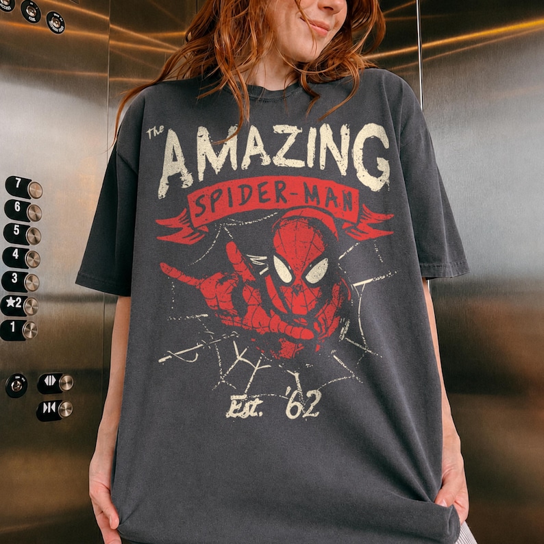 May include: A charcoal gray short-sleeve t-shirt featuring a distressed graphic of Spider-Man in red and white. The text reads The AMAZING SPIDER-MAN and Est. '62, with a spiderweb design integrated into the background of the central character illustration.
