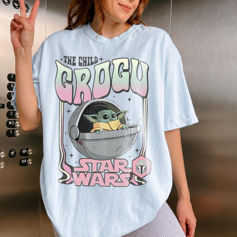 K&ouml;nnte beinhalten: Ein hellblaues T-Shirt mit einem Grafikdruck von Grogu, auch bekannt als Das Kind, der in einer schwebenden Kapsel sitzt. Das Design enth&auml;lt den Text THE CHILD GROGU und STAR WARS in einer stilisierten Schrift, akzentuiert mit kleinen Sternen und einem Helm-Symbol.
