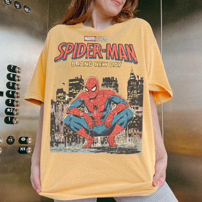 May include: A mustard yellow short-sleeve t-shirt featuring a graphic print of Spider-Man crouching in front of a city skyline. The text reads Marvel Studios Spider-Man Brand New Day. The design has a distressed, vintage-inspired aesthetic.