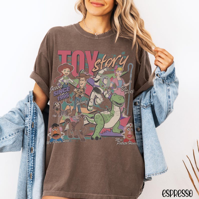 May include: Espresso-colored t-shirt featuring a graphic of Toy Story characters, including Woody, Buzz Lightyear, and Jessie. The design includes the text "Toy Story" and "Jessie Since 1995". The shirt has a vintage, worn look.