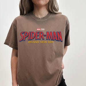 May include: A brown short-sleeved t-shirt featuring the Marvel Studios logo above the red and blue text Spider-Man and the yellow text Brand New Day printed on the chest. The shirt has a crew neckline and a standard fit design.