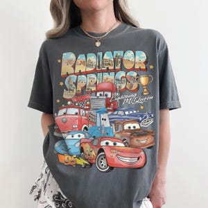 Op de afbeelding: Een antracietgrijs T-shirt met korte mouwen en een grafische print van Radiator Springs uit de film Cars. Het ontwerp bevat Lightning McQueen, Mater en andere personages, samen met de tekst Radiator Springs en Lightning McQueen.