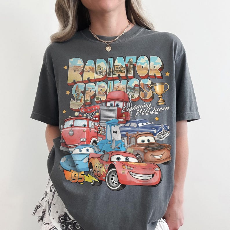 Op de afbeelding: Een antracietgrijs T-shirt met korte mouwen en een grafische print van Radiator Springs uit de film Cars. Het ontwerp bevat Lightning McQueen, Mater en andere personages, samen met de tekst Radiator Springs en Lightning McQueen.