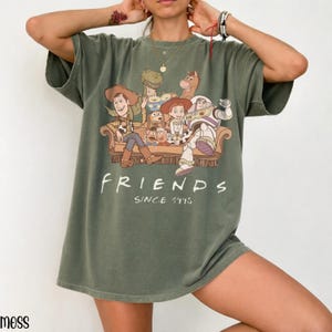 May include: An olive green t-shirt with a graphic of Toy Story characters on a sofa, with the word "FRIENDS" and "SINCE 1995" printed below. The shirt has short sleeves and a relaxed fit.