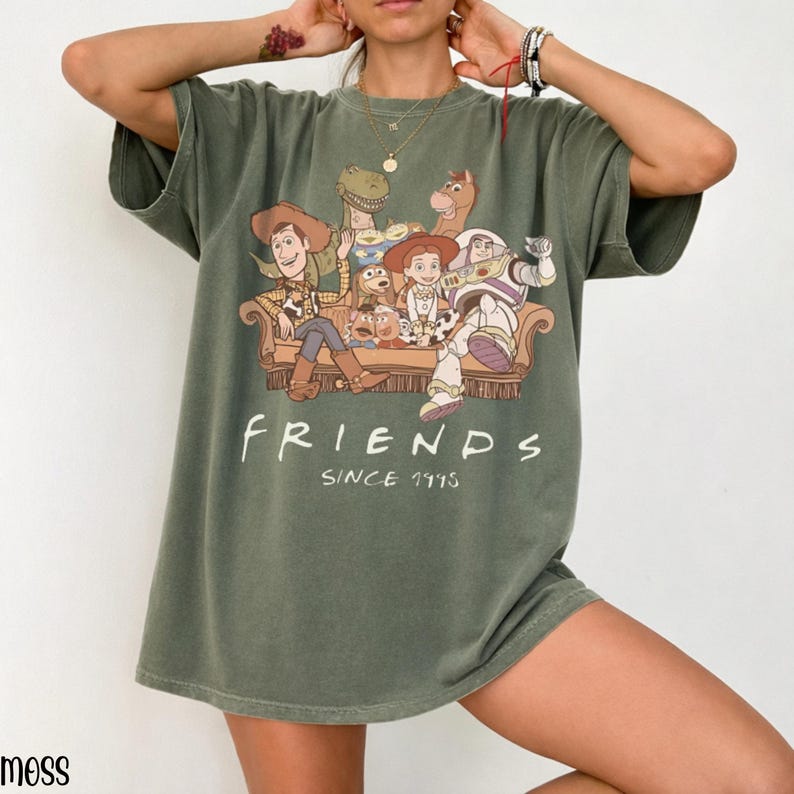 May include: An olive green t-shirt with a graphic of Toy Story characters on a sofa, with the word "FRIENDS" and "SINCE 1995" printed below. The shirt has short sleeves and a relaxed fit.
