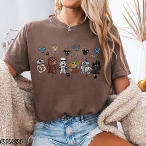 May include: Espresso-colored t-shirt featuring a graphic of Star Wars characters holding balloons. The characters include BB-8, Chewbacca, a Stormtrooper, Grogu, R2-D2, and Darth Vader. The balloons have various designs, and the shirt has a relaxed fit.
