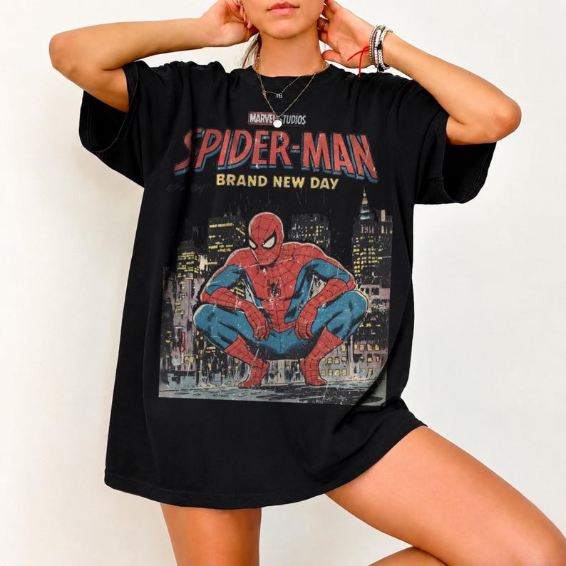 May include: A black short-sleeve t-shirt featuring a graphic print of the Marvel Studios Spider-Man character in a crouching pose. The text above the character reads "SPIDER-MAN BRAND NEW DAY" in red and yellow lettering against a city skyline.