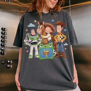 May include: Dark gray t-shirt featuring a graphic of characters from Toy Story 5. The design includes Buzz Lightyear, Woody, Jessie, and a horse, with the movie title on a green sign. Stars and clouds are also included.