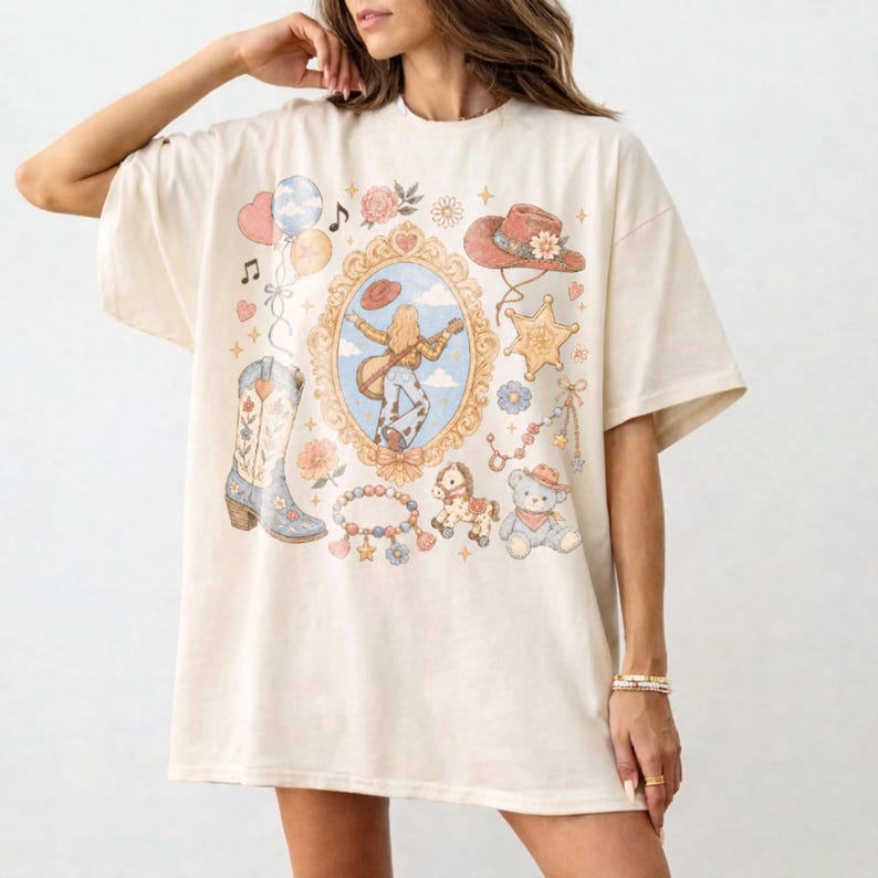 Puede incluir: Una camiseta de manga corta color hueso con un gr&aacute;fico central de espejo ovalado que muestra a una persona con una guitarra. Los elementos incluyen una bota vaquera, un sombrero, una estrella de sheriff, un oso de peluche, un caballo y flores.