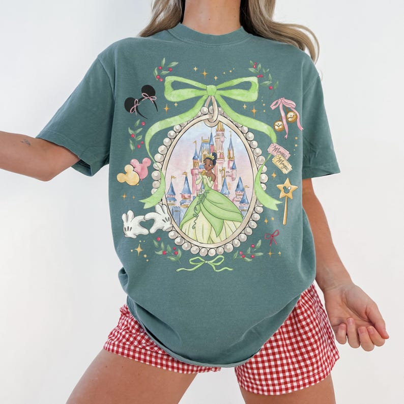 Pu&ograve; includere: T-shirt color verde acqua con una principessa in abito verde all'interno di una cornice ovale, con un castello sullo sfondo. Il design include un fiocco verde, orecchie di Topolino e una bacchetta magica. La modella indossa pantaloncini a quadri rossi e bianchi.