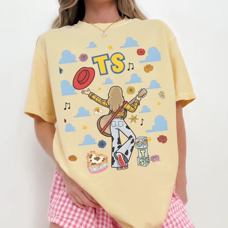 May include: A pale yellow t-shirt featuring a graphic illustration of a person with long blonde hair holding a guitar and a red hat. The design includes blue clouds, yellow stars, musical notes, flowers, and blocks with the letters T and S.