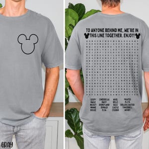 May include: Gray t-shirt featuring a black Mickey Mouse outline on the front. The back of the shirt has the text "TO ANYONE BEHIND ME, WE'RE IN THIS LINE TOGETHER. ENJOY!" with a word search of Disney characters.