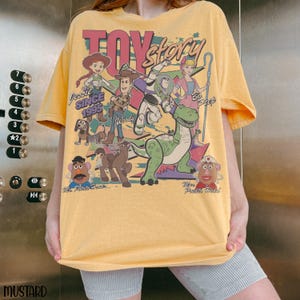 May include: Mustard yellow t-shirt featuring a graphic of Toy Story characters, including Woody, Buzz Lightyear, and Jessie. The text "TOY STORY" is at the top, with "SINCE 1995" below Jessie. The shirt has a vintage, distressed look.