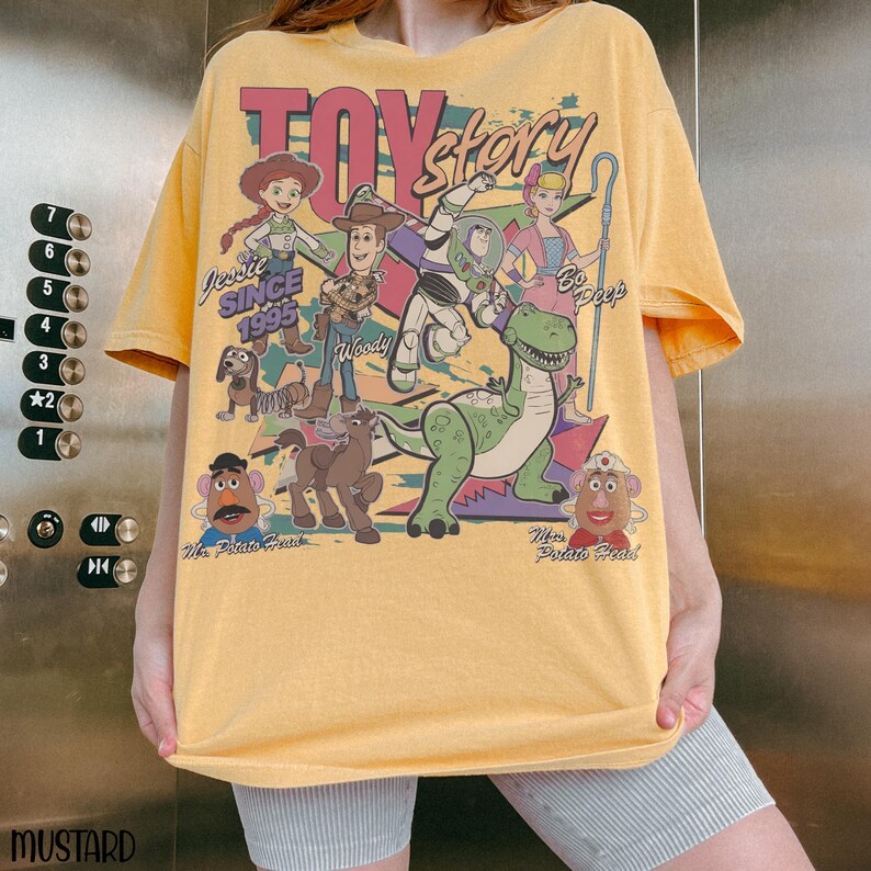 May include: Mustard yellow t-shirt featuring a graphic of Toy Story characters, including Woody, Buzz Lightyear, and Jessie. The text "TOY STORY" is at the top, with "SINCE 1995" below Jessie. The shirt has a vintage, distressed look.