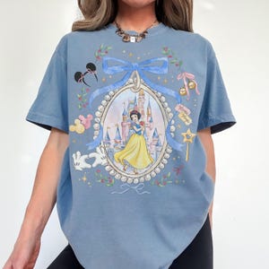 Pu&ograve; includere: T-shirt azzurra con un'illustrazione incorniciata di Biancaneve davanti a un castello. La cornice &egrave; decorata con un nastro blu, perle e icone a tema Disney. La maglietta ha maniche corte.