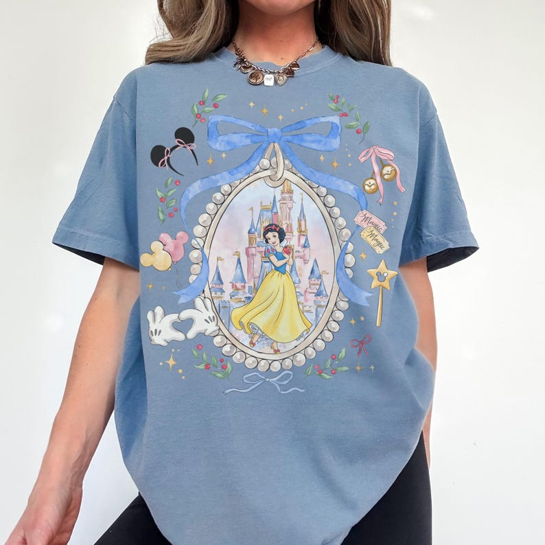 Pu&ograve; includere: T-shirt azzurra con un'illustrazione incorniciata di Biancaneve davanti a un castello. La cornice &egrave; decorata con un nastro blu, perle e icone a tema Disney. La maglietta ha maniche corte.