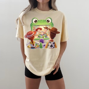 Puede incluir: Camiseta amarillo claro con un gr&aacute;fico colorido de personajes de la pel&iacute;cula Toy Story. El dise&ntilde;o muestra a Woody, Buzz Lightyear, Jessie y otros personajes dentro de un marco verde con forma de rana. La camiseta es de estilo casual, de manga corta.