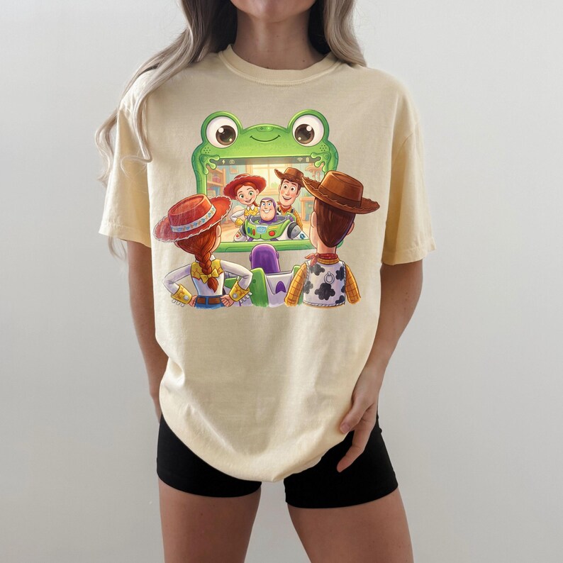 Puede incluir: Camiseta amarillo claro con un gr&aacute;fico colorido de personajes de la pel&iacute;cula Toy Story. El dise&ntilde;o muestra a Woody, Buzz Lightyear, Jessie y otros personajes dentro de un marco verde con forma de rana. La camiseta es de estilo casual, de manga corta.