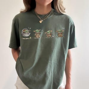 May include: An olive green short-sleeve t-shirt featuring a graphic print of a small green creature in four different poses. The illustrations show the character in a floating pod, using the force, sitting with a cup, and waving with the text "coo!"