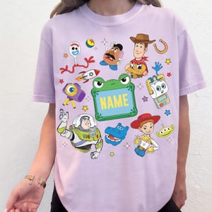 May include: A light purple t-shirt featuring a colorful graphic print of various animated characters including a cowboy, a space ranger, a cowgirl, a potato figure, and a fork character, with a central green frog frame containing the word NAME.