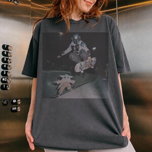 Pu&ograve; includere: Una t-shirt a maniche corte color grigio antracite con una stampa grafica raffigurante una figura con casco e armatura che esegue un trick con lo skateboard in aria sopra una piccola creatura dalle grandi orecchie su uno skateboard di notte.