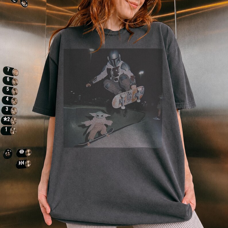 Pu&ograve; includere: Una t-shirt a maniche corte color grigio antracite con una stampa grafica raffigurante una figura con casco e armatura che esegue un trick con lo skateboard in aria sopra una piccola creatura dalle grandi orecchie su uno skateboard di notte.