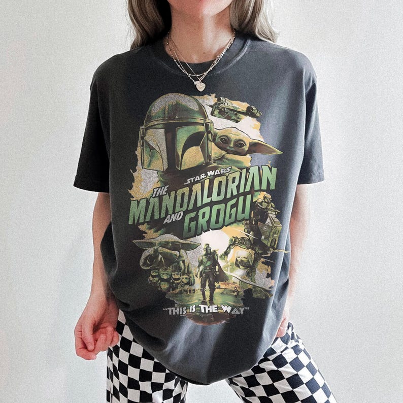 May include: Dark gray t-shirt featuring a graphic from the Star Wars series, "The Mandalorian and Grogu." The design includes images of the Mandalorian, Grogu, and other characters, with the phrase "This is the Way" at the bottom.