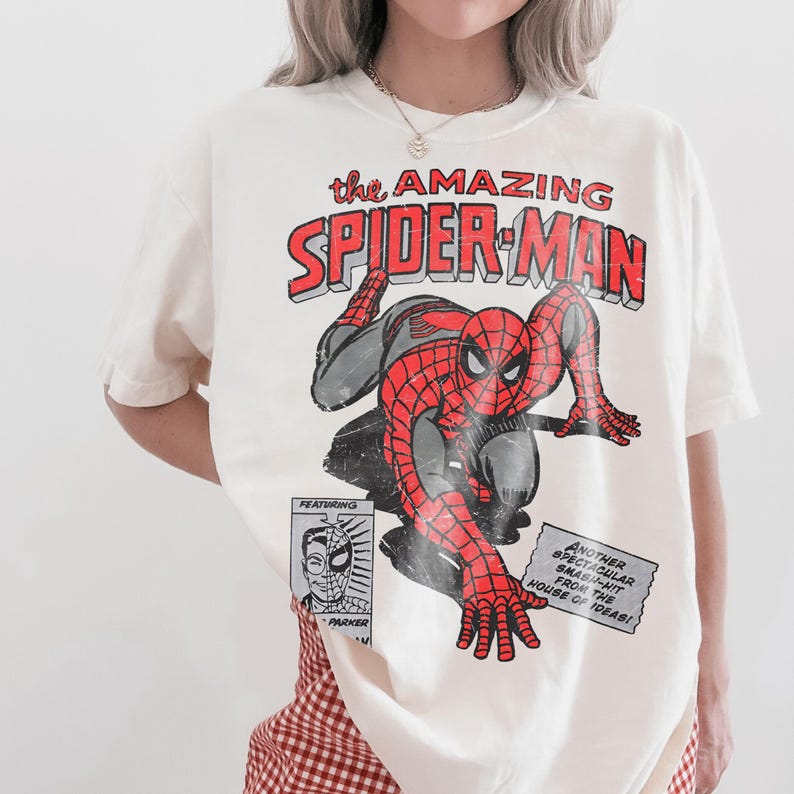 May include: An off-white short-sleeve t-shirt featuring a distressed graphic of The Amazing Spider-Man in red and black. The print includes the text 'the AMAZING SPIDER-MAN', a small portrait of Parker, and a box reading 'ANOTHER SPECTACULAR SMASH-HIT FROM THE HOUSE OF IDEAS!'
