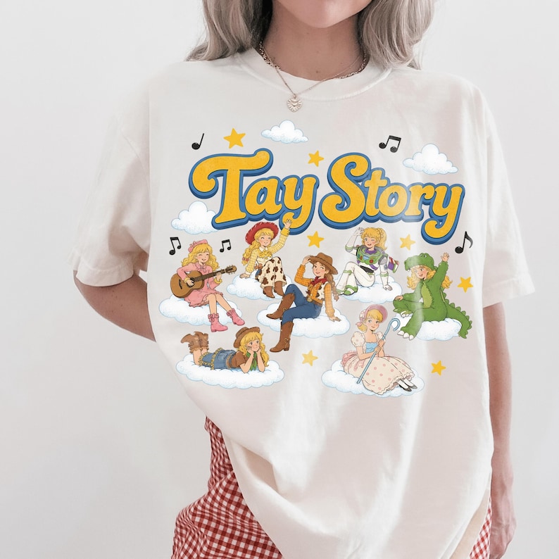 Puede incluir: Una camiseta de manga corta de color crema con el texto "Tay Story" en letras amarillas grandes. El gr&aacute;fico incluye varios personajes ilustrados con diversos atuendos, cada uno sentado sobre una nube blanca, rodeado de estrellas amarillas y notas musicales negras.