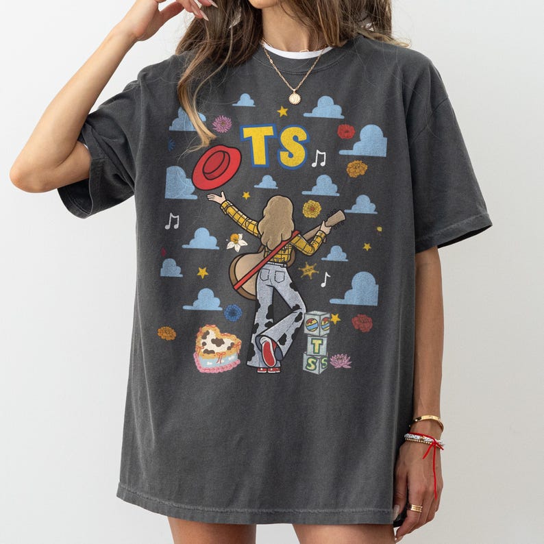 May include: A charcoal grey short-sleeve t-shirt featuring a graphic print of a person with long hair playing a guitar, wearing a yellow plaid shirt and cow-print trousers. The design includes a red hat, blue clouds, yellow stars, and the text TS.
