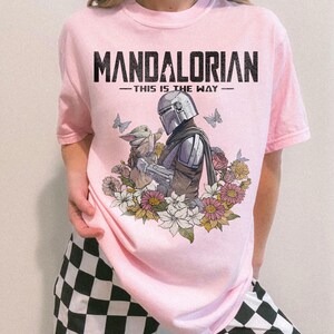 May include: Pink t-shirt featuring a graphic of The Mandalorian holding Grogu, surrounded by flowers and butterflies. The text "MANDALORIAN - THIS IS THE WAY -" is printed across the top in black.