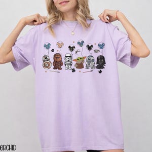 May include: A light purple t-shirt featuring cartoon Star Wars characters holding Mickey Mouse balloons. The characters include BB-8, Chewbacca, a Stormtrooper, Grogu, R2-D2, and Darth Vader. The word "ORCHID" is printed at the bottom.