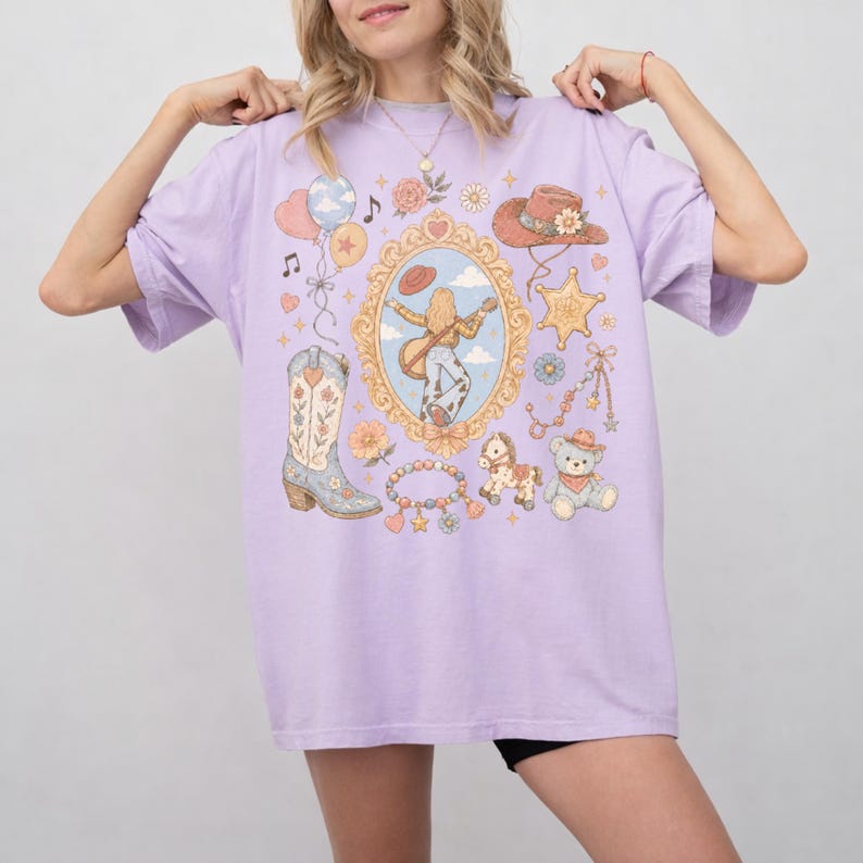 Puede incluir: Una camiseta de color morado claro con un gr&aacute;fico central de una persona tocando la guitarra dentro de un marco dorado ornamentado. Alrededor del marco hay ilustraciones de una bota vaquera, un sombrero, una insignia de estrella y flores.