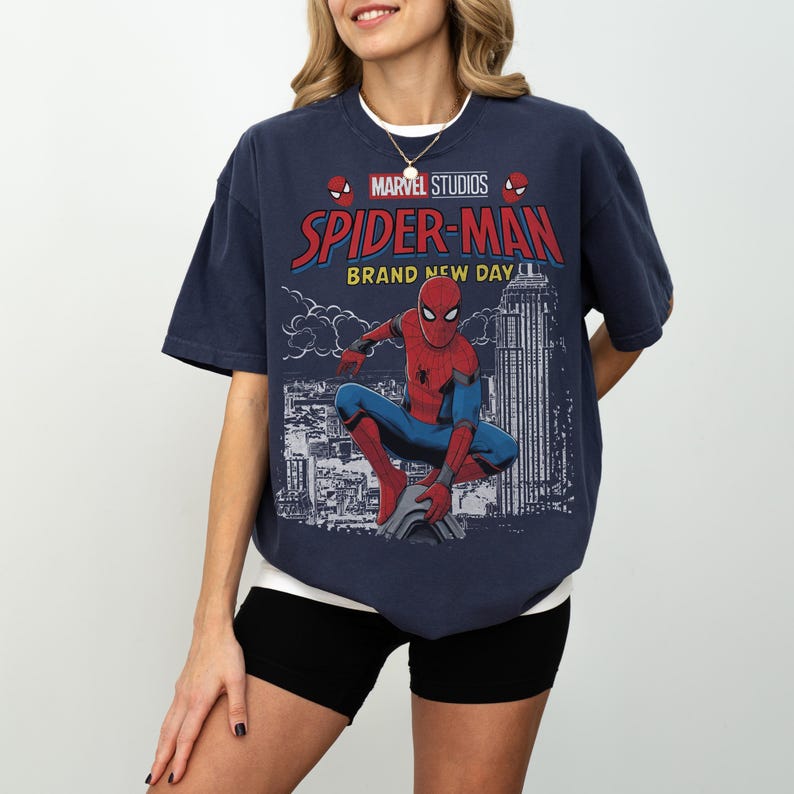 May include: A dark blue short-sleeve t-shirt featuring a graphic print of Spider-Man crouching on a building. The text reads Marvel Studios, Spider-Man, and Brand New Day. The design includes a cityscape background with clouds and a tall skyscraper.
