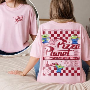 May include: A light pink t-shirt featuring the "Pizza Planet" logo on the front and back. The back design includes the text "Pizza Planet", aliens, and the phrase "Friday Night Kid Night". The shirt is a casual, oversized fit.