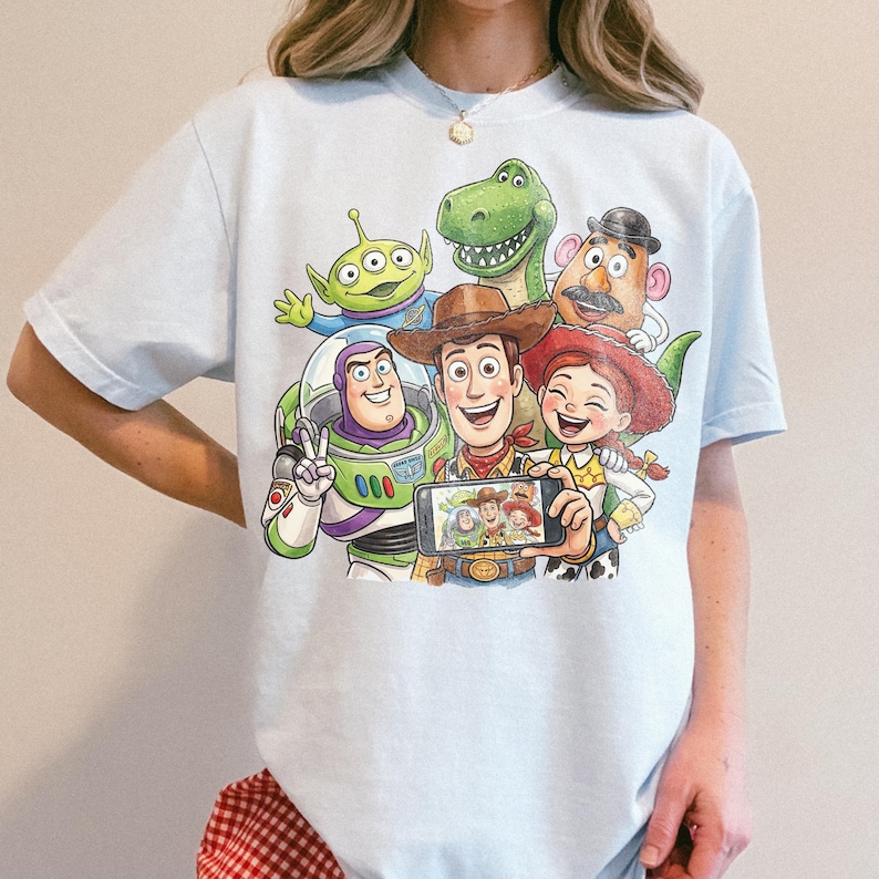 May include: Light blue t-shirt featuring a colorful graphic of Toy Story characters taking a selfie. The design includes Woody, Buzz Lightyear, Rex, Mr. Potato Head, Jessie, and an alien. The shirt is a comfortable, casual style.